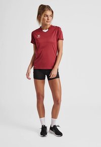 Rotes Sport-T-Shirt mit V-Ausschnitt, kurzen Ärmeln und Logo. Kombiniert mit schwarzen Shorts und schwarzen Sneakers, dazu weiße Knöchelsocken.