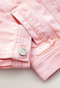 Zbliżenie na mankiet jasnoróżowej kurtki jeansowej z srebrnym zatrzaskiem i widocznym przeszyciem szwu kieszeni.