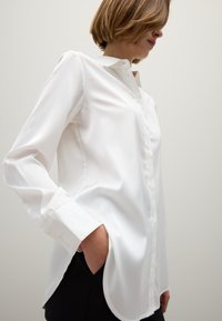 Camisa blanca de manga larga con botones, cuello clásico, textura suave y dobladillo redondeado, llevada sobre una prenda negra.