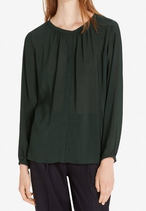 Vrouw die een donkergroene blouse met lange mouwen, een ronde hals en subtiele plooien draagt, gecombineerd met een zwarte broek.