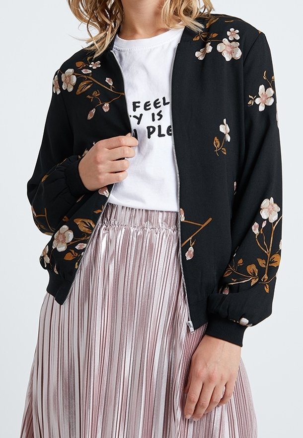 Femme portant une veste zippée noire à fleurs, un t-shirt blanc imprimé et une jupe plissée métallique, tenant la fermeture éclair de la veste près du col.