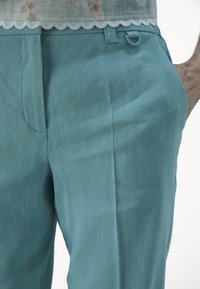 Teollanssia pellavahousuja, joissa on sileä rakenne, sivutaskut ja vyötäröstä roikkuva yksityiskohta. Yhtenäinen väri, jossa on hieno kiilto.