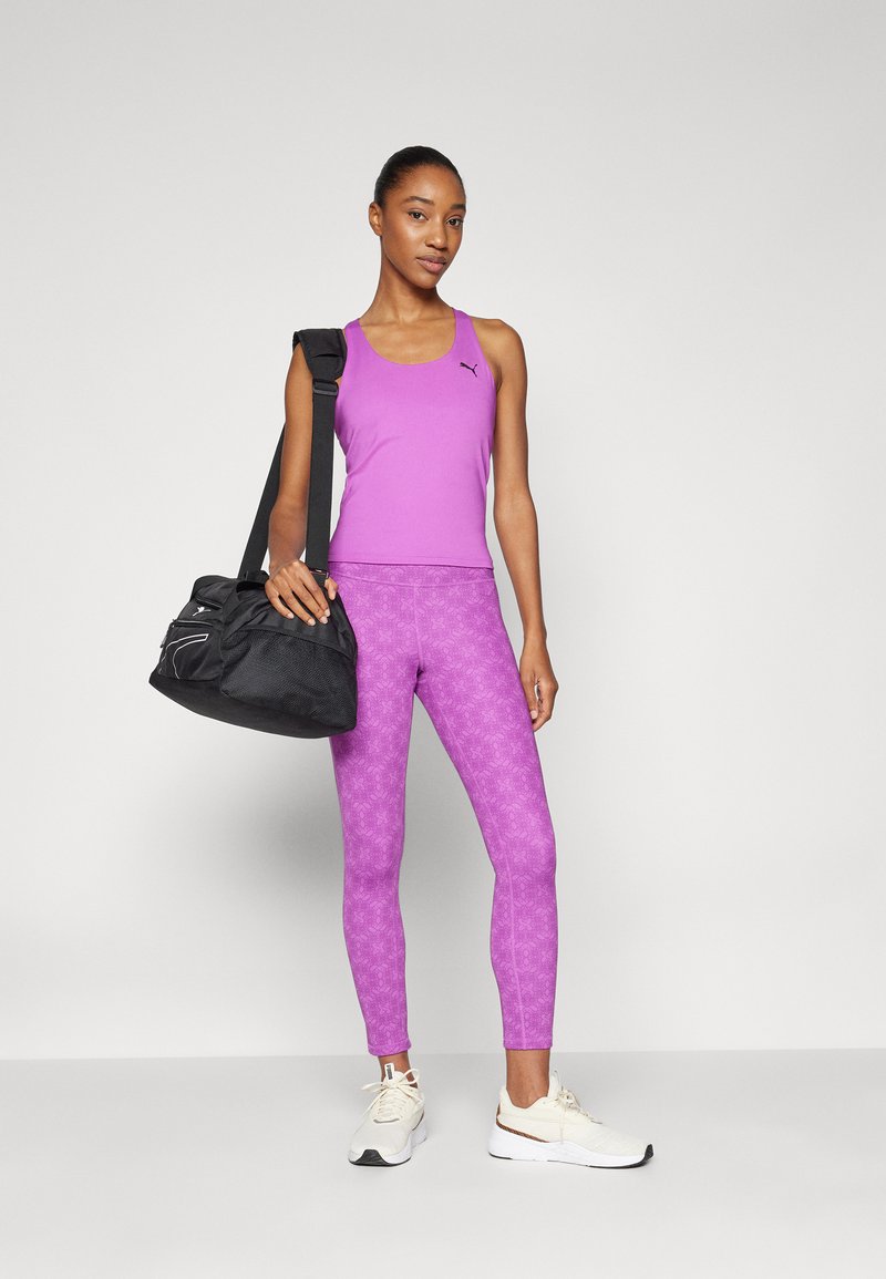 Lila Sporttop und gemusterte Leggings, kombiniert mit einer schwarzen Sporttasche. Weiße Sportschuhe vervollständigen das Ensemble.