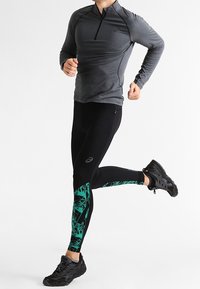 Leggings de sport noirs avec un motif géométrique vert sur le bas des jambes, associés à un haut ajusté gris à manches longues avec une fermeture éclair à moitié. Pose de course.