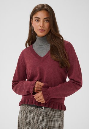 Frau mit langen braunen Haaren, die einen violetten Pullover mit V-Ausschnitt über einem grauen Rollkragenpullover und einem karierten Rock trägt und vor einem schlichten Hintergrund steht.