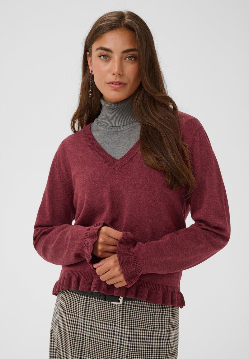 Femme aux longs cheveux bruns portant un pull violet à col en V par-dessus un col roulé gris et une jupe à carreaux, debout devant un fond uni.