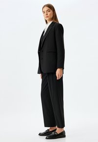 Costume noir sur mesure avec un blazer à un bouton et un pantalon à jambes larges, associé à des mocassins noirs. Tissu lisse, design minimaliste.
