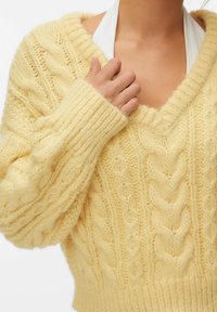 Personne portant un pull à torsades épais de couleur jaune pâle à manches longues, tenant l'encolure d'une main, un haut blanc visible en dessous.