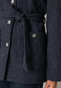 Granatowy płaszcz z tweedu z paskiem w talii, ozdobiony srebrzystymi guzikami w kształcie węzłów oraz przednią kieszenią, wykonany z teksturowanej tkaniny.