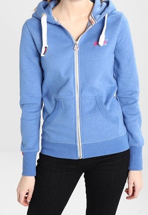 Woman wearing a light blue zip-up hoodie with white drawstrings and black pants, standing against a plain gray background.