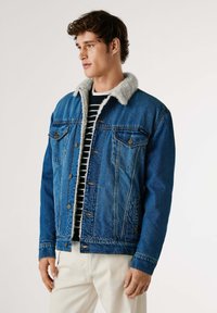Denimjacka i blått med en mjuk, grå pälskrage. Har två bröstfickor och knäppning med knappar, bärs över en randig svart och vit topp.
