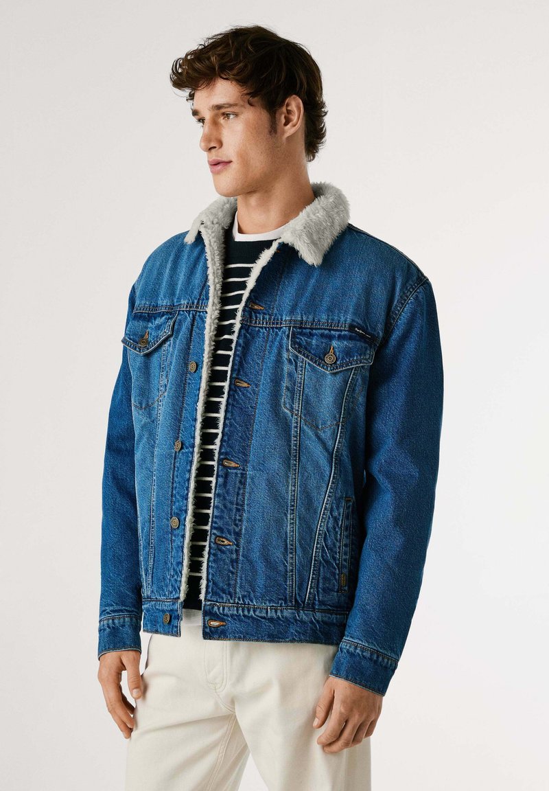 Denimjacka i blått med en mjuk, grå pälskrage. Har två bröstfickor och knäppning med knappar, bärs över en randig svart och vit topp.