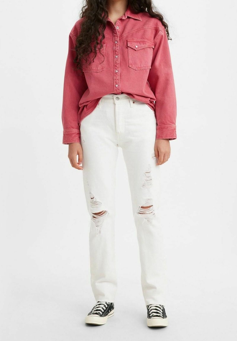 Femme portant une chemise rouge délavée à boutons, un jean blanc déchiré et des baskets basses noires, debout devant un fond blanc uni.
