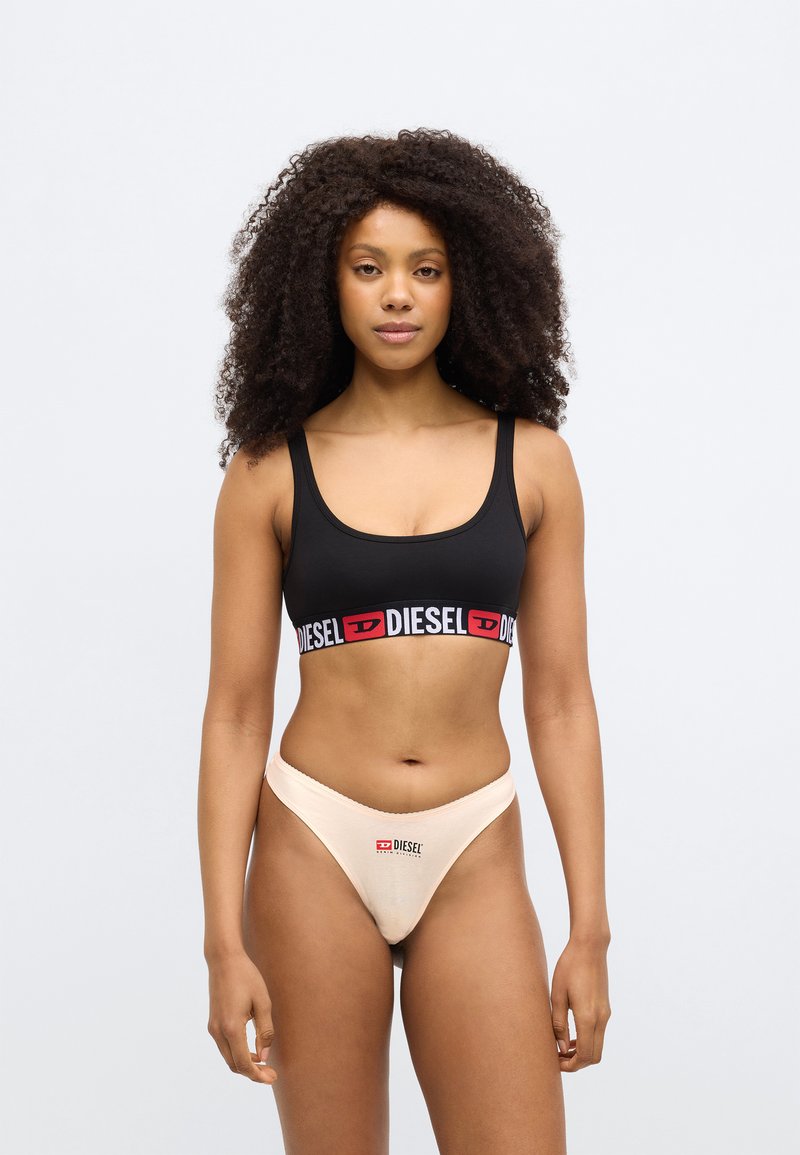 Black sports bra with red and white "DIESEL" logo band; paired with beige thong featuring a similar logo. Smooth fabric texture throughout.