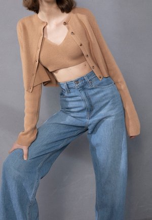 Kvinde iført en kort camelfarvet cardigan og matchende strikket brystholder, kombineret med højtaljede, vidde bukser i lys blå denim, poserer mod en grå baggrund.