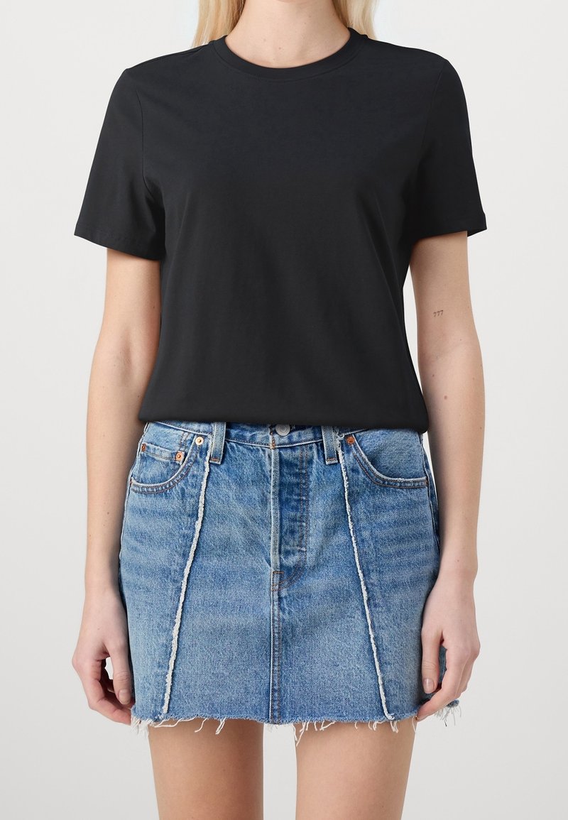 Personne blonde portant un t-shirt noir uni à manches courtes rentré dans une mini-jupe en denim bleu avec un ourlet effiloché et des coutures verticales.