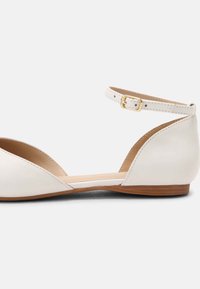 Bailarinas de cuero blanco con un tacón bajo, lados abiertos y una correa para el tobillo con un detalle de hebilla dorada. Textura suave y diseño minimalista.