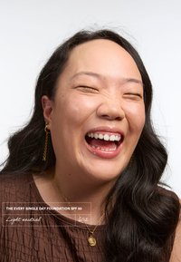 Botella de base etiquetada "The Every Single Day Foundation SPF 50, Light Neutral 602" frente a una persona sonriente con cabello largo y negro.