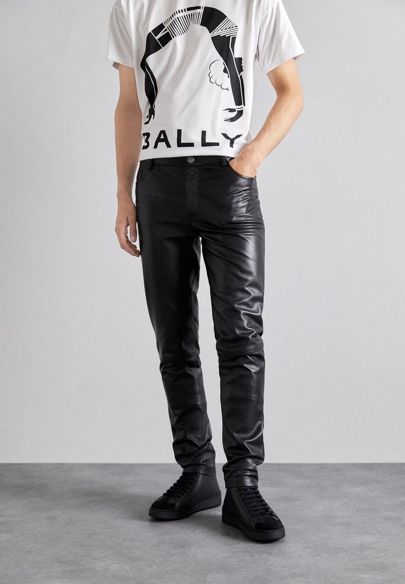 Svarta läderbyxor med smal passform och glans, kombinerat med en vit T-shirt med svart grafisk design och texten "BALLY." Svarta högskräpade sneakers.