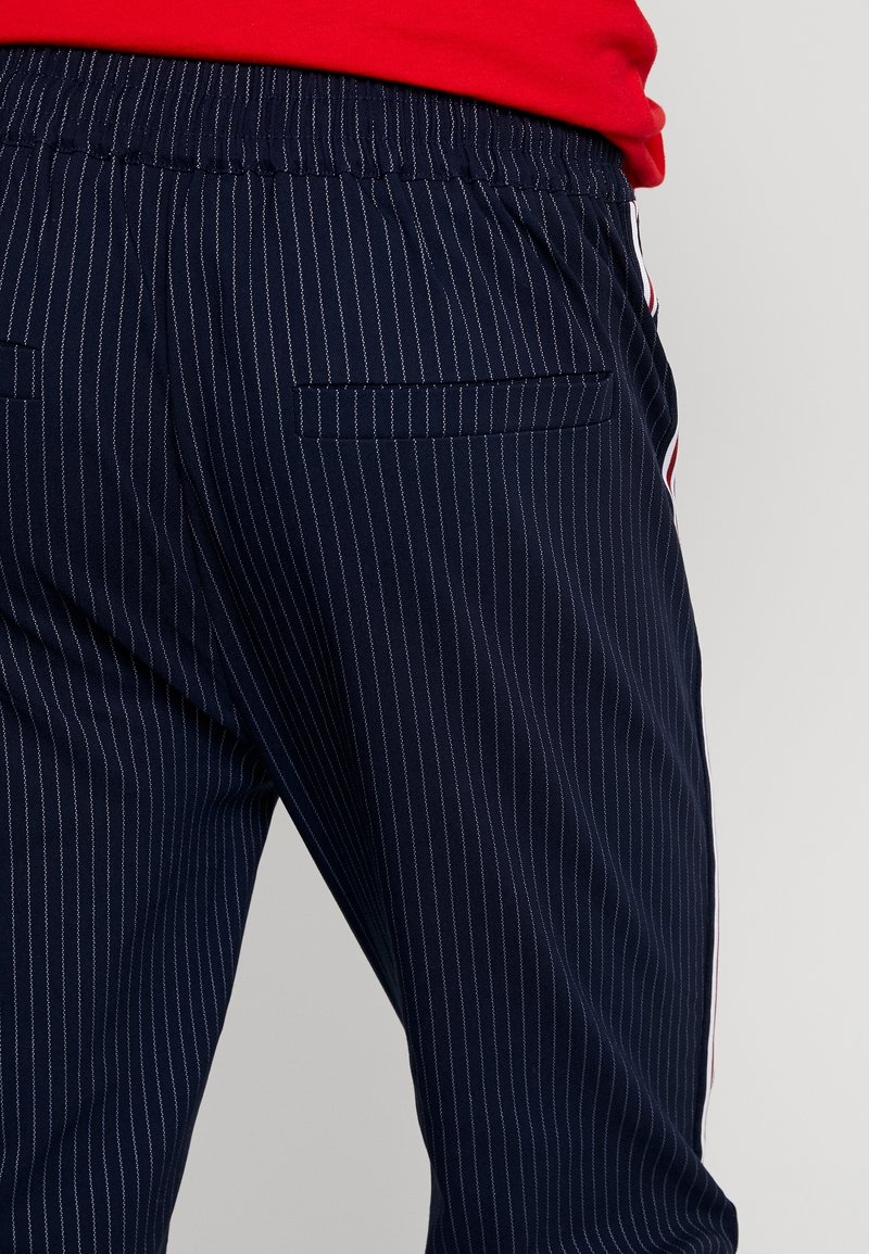 Pantalon bleu marine à fines rayures, avec taille élastique, présentant deux poches arrière et des bandes latérales contrastantes en rouge et blanc.