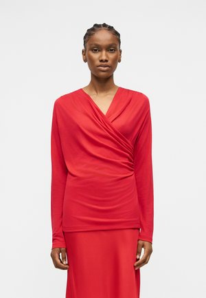 Žena s pletenými vlasmi, oblečená v červenom dlhom rukáve, zavinovacom topu a zodpovedajúcej červenej sukni, stojaca na jednoduchom svetlom pozadí.