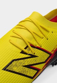 Κίτρινο αθλητικό παπούτσι με κίτρινες κορδόνια και μαύρο και κόκκινο στυλιζαρισμένο λογότυπο "N" στο υφασμάτινο πάνω μέρος.