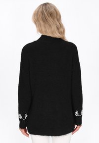 Czarny, oversize sweter z puszystego materiału, z długimi rękawami i białymi haftowanymi akcentami na mankietach. Prosty, minimalistyczny design.