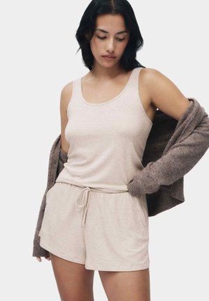 Kvinde med mørkt hår iført beige ærmeløs top og matchende shorts, der trækker en brun cardigan af med højre hånd.