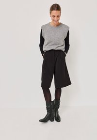 Femeie îmbrăcată într-un vest gri fără mâneci peste un top lung cu mânecă neagră, pantaloni scurți largi negri, colanți negri și cizme negre până la jumătatea gambei, stând în picioare și zâmbind.