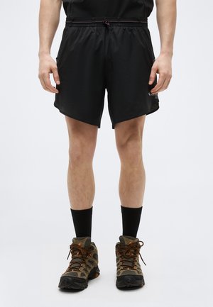 Muž oblečený v čiernych športových šortkách, čiernych ponožkách po členky a hnedej turistickej obuvi, stojaci s uvoľnenými rukami pri tele.