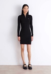 Rochie neagră cu dungi, cu mâneci lungi și guler cu fermoar, cu accente roșii la manșete și margine. Modelul poartă pantofi plata negri.