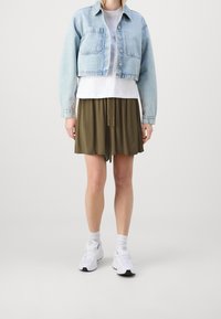 Giacca di jeans corta azzurro chiaro, t-shirt bianca, gonna plissettata verde oliva e sneakers bianche con dettagli grigi, il tutto su uno sfondo neutro.