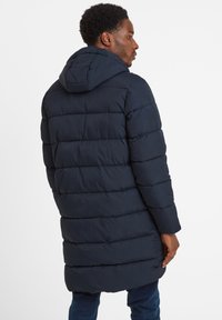 Man seen from the back wearing a long navy blue padded jacket with horizontal quilting and a hood, paired with blue jeans.