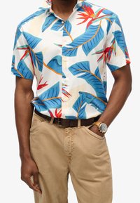 Camisa de manga corta en tela ligera con base blanca, decorada con hojas azules y flores rojas. Combinada con pantalones beiges y un cinturón marrón.