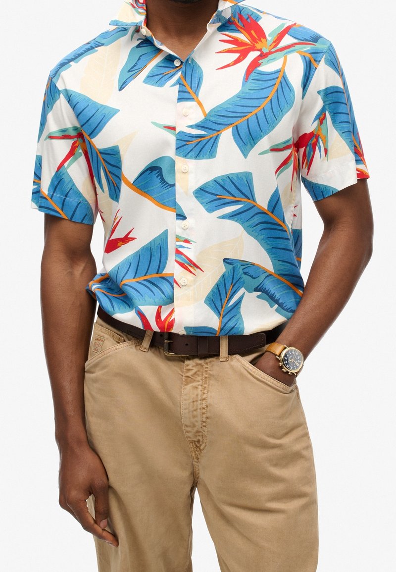 Camisa de manga corta en tela ligera con base blanca, decorada con hojas azules y flores rojas. Combinada con pantalones beiges y un cinturón marrón.