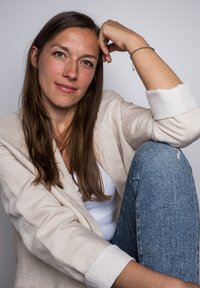 Bézs vászonblézer egy fehér felsőn, kék, koptatott farmerral párosítva. Hosszú, barna haj és egy ezüst karkötő emeli a megjelenést. Semleges háttér.