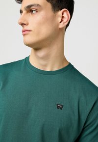 Grön bomullströja med ribbat rundhals och en liten svart Wrangler-logotyp på vänster bröstområde. Slät, lätt textur.