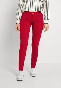 Mujer vistiendo unos pantalones skinny rojos brillantes, zapatillas blancas con cordones y un suéter corto blanco con rayas verticales negras.