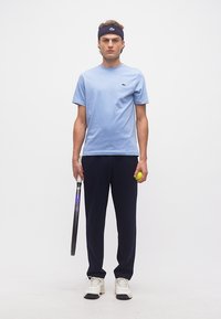 Άνδρας στέκεται κρατώντας ρακέτα τένις στο δεξί χέρι και μπάλα τένις στο αριστερό, φοράει ανοιχτό μπλε μπλουζάκι, σκούρο μπλε παντελόνι, λευκά αθλητικά παπούτσια και σκούφο κεφαλής σκούρου μπλε χρώματος.