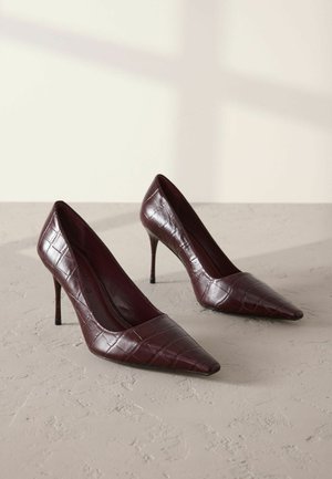 Pump bordeaux con tacco alto, punta affusolata, texture lucida a croco e tacco sottile. Design classico con accenti minimi.