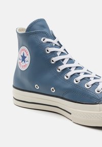 Kék bőr magasszárú sneaker fehér fűzővel. Jellemzője egy kerek logó folt, fém fűzőlyukak és egy texturált fehér gumitalp.