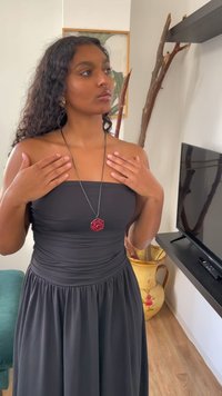 Robe noire sans bretelles avec un tissu froncé. Un pendentif en rose rouge sur un collier en corde noire. Cheveux bouclés et mains reposant sur les épaules. Accents en bois visibles.
