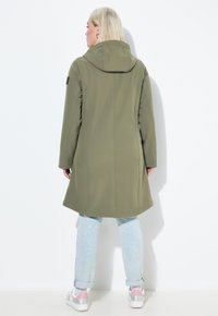 Olivově zelený, dlouhý kabát vyrobený z měkkého materiálu, má kapuci, boční kapsy a rovný střih, v kombinaci se světle modrými džíny a teniskami.