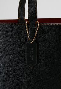 Czarna torebka tote z teksturowanej skóry o strukturalnym kształcie, kontrastowe przeszycia oraz złoty łańcuszek przymocowany do prostokątnej zawieszki.