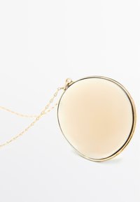 Collana oro con un pendente rotondo, liscio e beige, con un sottile bordo metallico e una delicata catena. Il design è minimalista ed elegante.