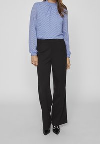 Blouse bleu à motifs, à manches longues avec un col froncé, associée à un pantalon noir à jambes larges et des chaussures noires à talons pointus.