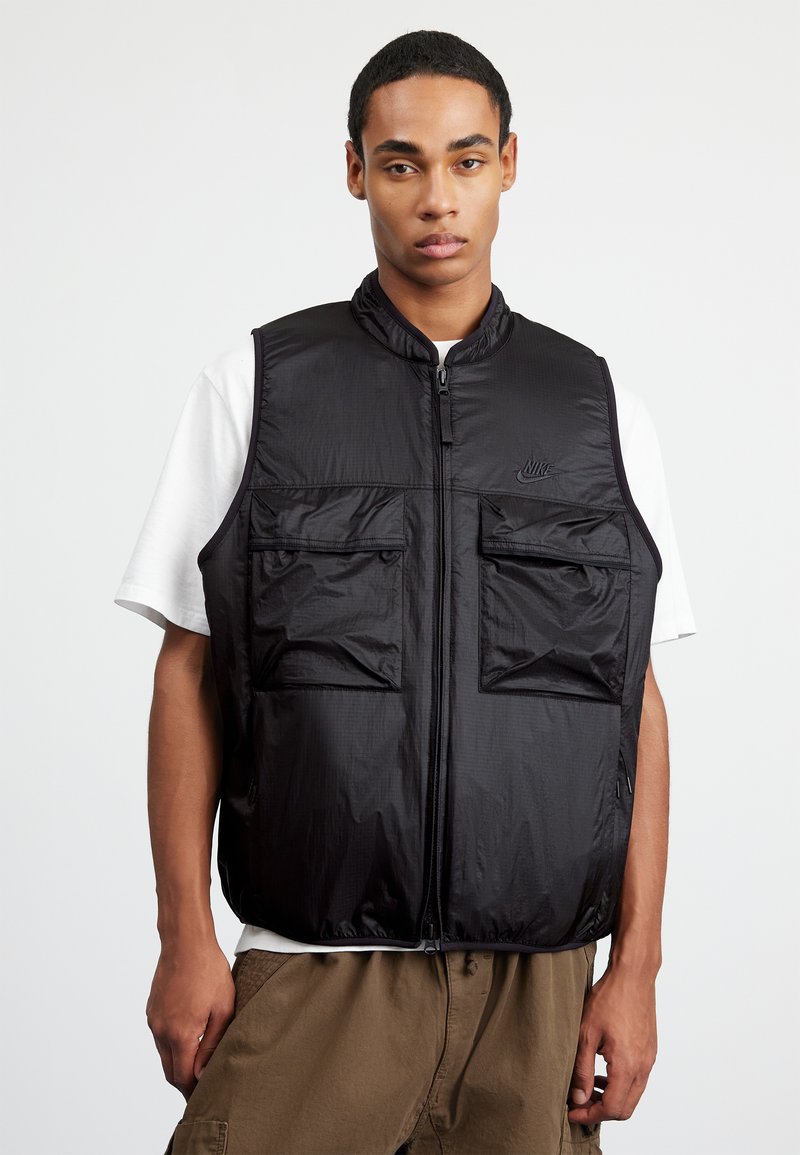 Μαύρο νάιλον γιλέκο με υψηλό κολάρο, κλείσιμο με φερμουάρ και δύο μπροστινές τσέπες. Φοριέται πάνω από λευκό πουκάμισο, το οποίο έχει λογότυπο στο στήθος.