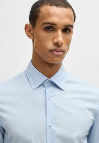Camicia azzurro chiaro con colletto a punta strutturato, chiusura con bottoni e texture liscia, caratterizzata da un design aderente e una leggera lucentezza.