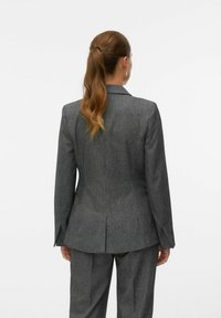Pielāgots pelēks žakets ar apaļkārtas rakstu, kas izceļ strukturētas plecus, vienu šķēlumu un šauru piegriezumu profesionālam izskatam.