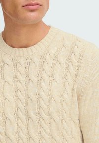 Pulover crem din tricot cu model cable-knit, având un decolteu rotund ribat și modele texturate de cabluri împletite pe întreaga suprafață.